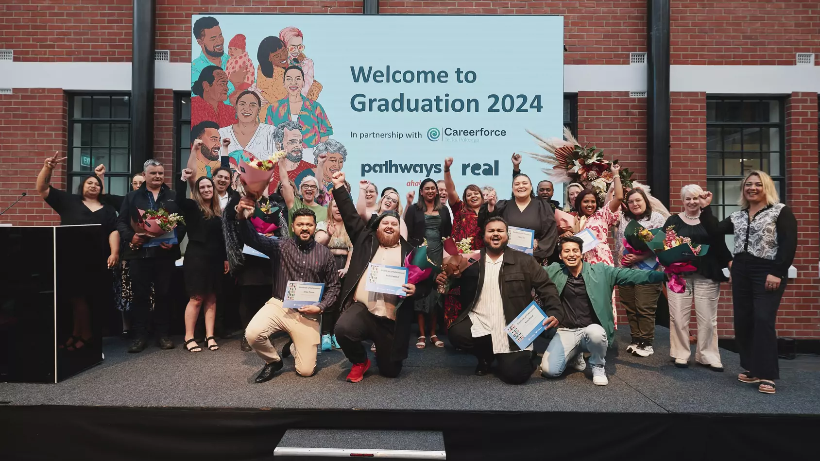 Graduates stand smiling and proud on stage with their certificates